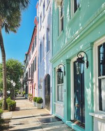 Sunlit pastel rowhouses (turquoise, lavender, peach) with black lanterns and a palm tree lining a charming historic southern coastal sidewalk under a bright blue sky