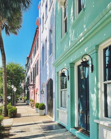 Sunlit pastel rowhouses (turquoise, lavender, peach) with black lanterns and a palm tree lining a charming historic southern coastal sidewalk under a bright blue sky