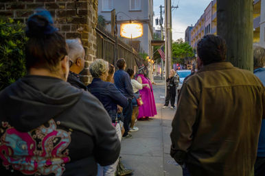 People queued along a downtown sidewalk at dusk beside a brick building and glowing sign, with a person in a bright pink dress and pointed hat, an American flag and urban storefronts in the background.