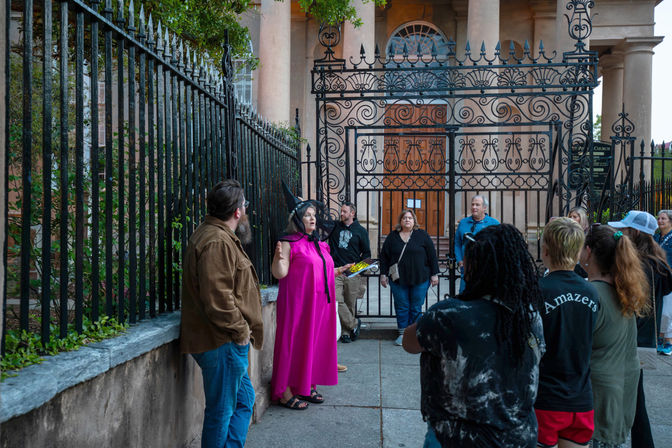 Tour guide in a bright magenta cape and pointed hat addresses a small tour group gathered outside an ornate wrought-iron gate of a historic church-like building