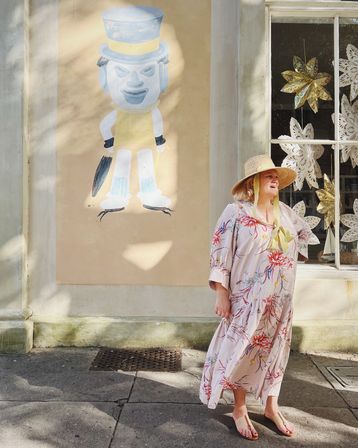 Sunlit street scene with a person in a wide-brim straw hat, ribbon, floral maxi dress and sandals standing on a sidewalk beside a pastel mural of a top-hatted cartoon and a shop window filled with paper star decorations.