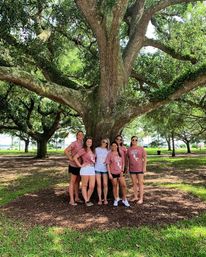 Six friends posing for a group photo under a sprawling oak tree in a sunny park, some wearing matching pink shirts and sunglasses, surrounded by green lawn and dappled shade.