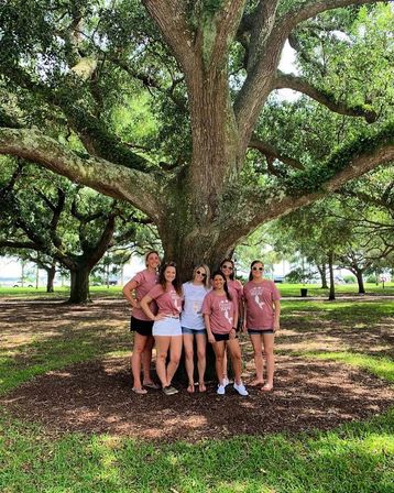 Six friends posing for a group photo under a sprawling oak tree in a sunny park, some wearing matching pink shirts and sunglasses, surrounded by green lawn and dappled shade.