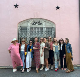 Group of friends in casual summer outfits posing and laughing in front of a pastel pink stucco wall with an arched window and decorative iron grille, some holding coffee cups — playful street photo.