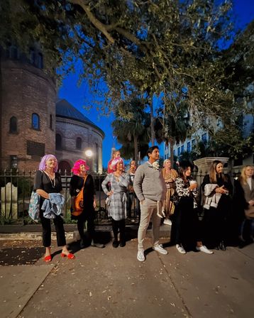 Evening crowd in colorful wigs and casual attire watching a street event outside a round historic brick church under oak trees and a glowing lamp.