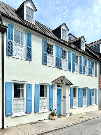 Sunlit historic townhouse with white stucco, powder-blue shutters and matching door, black metal roof with dormer windows, and a planter by the sidewalk.