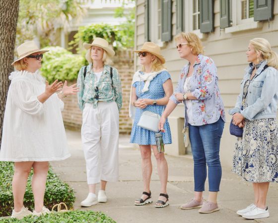 Group of five women on a guided walking tour along a tree-lined historic street, wearing sun hats and casual spring outfits while listening to a guide.