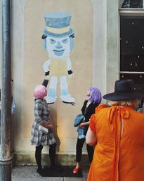 Three people in colorful wigs and outfits on a city sidewalk looking up at a playful blue-faced, top-hat mural painted on a pale wall next to a storefront window.