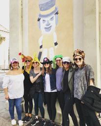 Seven women in playful hats and sunglasses pose on a city sidewalk in front of a large, cartoonish masked-figure mural painted on a beige building.