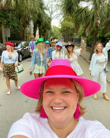 Smiling woman in a bright pink hat takes a selfie as friends in colorful hats stroll down a palm-lined residential street.