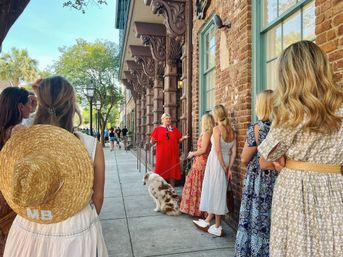Lively historic downtown walking tour: a guide in a red dress speaks to a group of women on a sunny sidewalk by brick buildings with ornate wooden corbels, lamp posts, trees and a leashed dog.