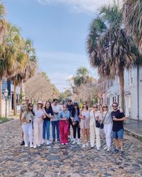 Group of friends posing on a sunny, palm-lined cobblestone street in a historic Southern coastal town, standing in front of pastel row houses and holding coffee cups.