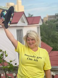 Smiling person on a rooftop deck wearing a yellow T-shirt that reads sounds gay i'm in, raising a black hat decorated with colorful stars, with red metal roofs, potted greenery and string lights in the background.