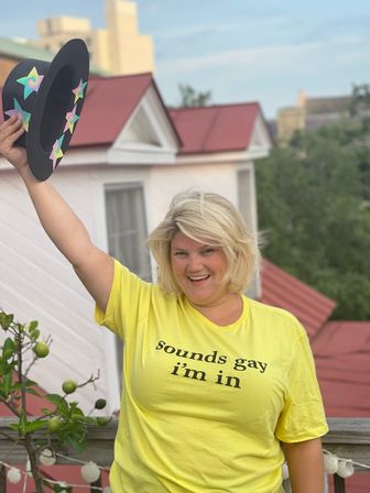 Smiling person on a rooftop deck wearing a yellow T-shirt that reads sounds gay i'm in, raising a black hat decorated with colorful stars, with red metal roofs, potted greenery and string lights in the background.