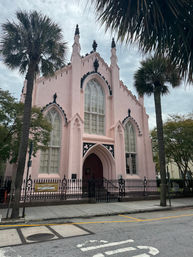 Pink Gothic Revival church with tall arched windows and decorative black trim, set behind a wrought-iron fence and flanked by palm trees on a southern coastal street.