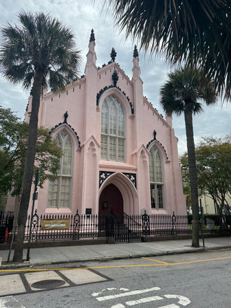 Pink Gothic Revival church with tall arched windows and decorative black trim, set behind a wrought-iron fence and flanked by palm trees on a southern coastal street.