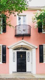 Cheerful pastel-pink townhouse facade with a glossy black paneled front door, white decorative entrance trim, small wrought-iron balcony above, black shutters and leafy green trees framing the scene.