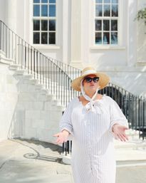 Person in a straw sun hat and sunglasses wearing a white striped summer dress, gesturing in front of a sunlit white neoclassical building with tall windows and a curved black iron staircase.