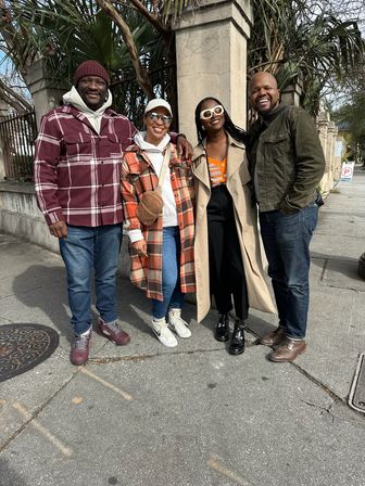 Four friends smiling and posing on a city sidewalk by stone pillars and palm trees, wearing cozy plaid jackets, a trench coat, jeans and sneakers.