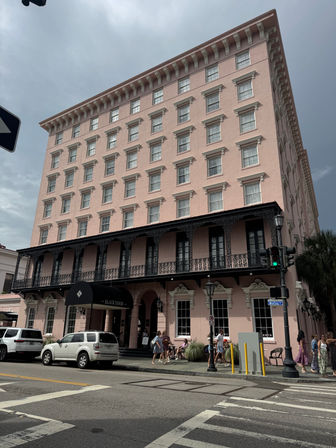 Pink historic hotel with rows of sash windows and ornate black wrought-iron balcony over a street-level entrance, parked cars and pedestrians at a downtown corner under an overcast sky.