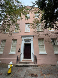 Charming pink three-story townhouse with white trim and dark front door, stone steps and black railings, yellow fire hydrant on the sidewalk and leafy branches framing the facade.