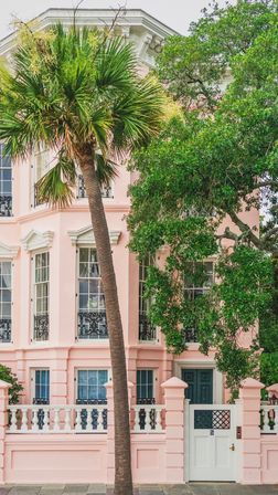 Pastel pink historic townhouse with tall palm tree, white balustrade and gate, decorative black wrought-iron balconies — coastal neighborhood charm.