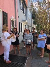 Guide leading a walking tour of women past pastel row houses with black shutters and flowering trees on a sunny historic downtown sidewalk