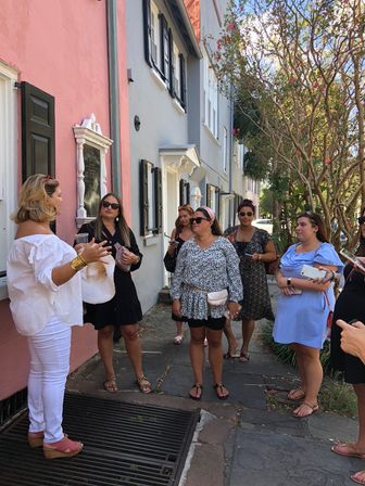 Guide leading a walking tour of women past pastel row houses with black shutters and flowering trees on a sunny historic downtown sidewalk