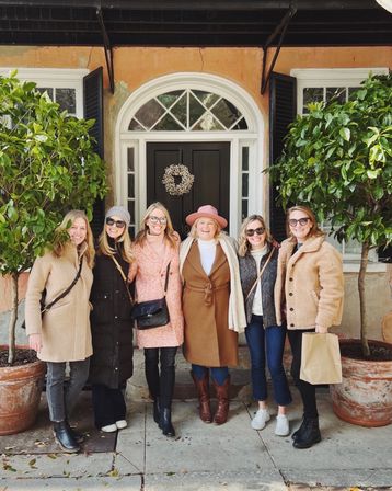 Group of six women in winter coats and boots smiling on a city sidewalk in front of a peach-colored historic storefront with black shutters, arched transom window, wreath and potted greenery.