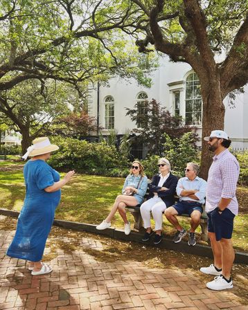 Tour guide in a straw hat speaking to four people seated on a bench beneath sprawling live oak trees in front of a historic white mansion in a sunlit garden setting.