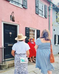 Three women in summer dresses and straw hats chatting in front of a pastel pink historic row house with black shutters and a dark door on a stone sidewalk — colorful sunny street scene in a charming historic district.