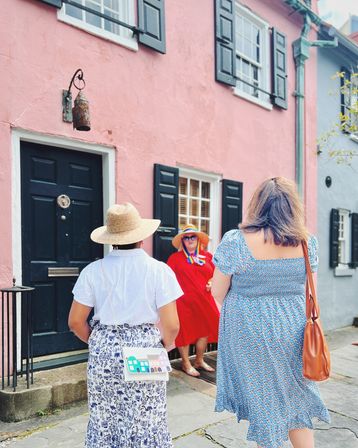 Three women in summer dresses and straw hats chatting in front of a pastel pink historic row house with black shutters and a dark door on a stone sidewalk — colorful sunny street scene in a charming historic district.