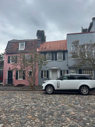 Quaint historic cobblestone street with a white SUV parked in front of pastel pink and blue-gray row houses, shuttered windows, tile roofs, bare trees and an overcast sky.