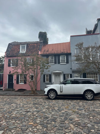 Quaint historic cobblestone street with a white SUV parked in front of pastel pink and blue-gray row houses, shuttered windows, tile roofs, bare trees and an overcast sky.