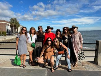 Group of smiling women in casual summer outfits and hats posing on a sunny seaside promenade by a seawall, with a palm tree, historic building and calm ocean under a blue sky