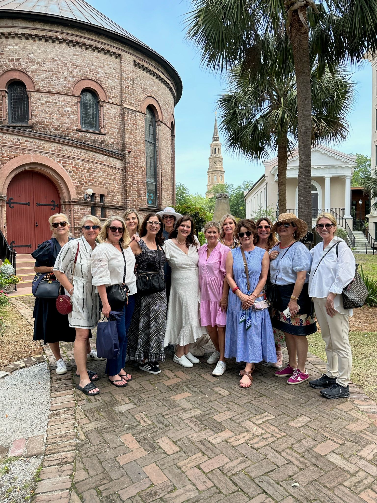 Smiling tour group of women posing in front of a round historic brick church with palm trees and a tall steeple visible in the background on a sunny day