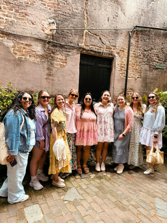 Cheerful group of friends in pastel summer dresses and sunglasses posing on a cobblestone alley in front of a weathered brick wall and black door.