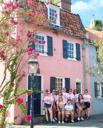 Bachelorette group in matching shirts posing outside a pastel pink historic townhouse with dark shutters, clay-tiled roof, blooming pink crepe myrtle and vintage street lamp.