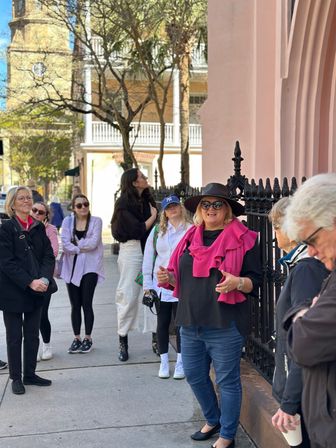 Tour guide in a wide-brim hat and bright pink sweater leading a small walking group on a historic downtown sidewalk beside a pink stucco building and ornate wrought-iron fence