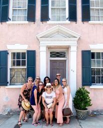 Eight women posing outside a pastel pink historic row house with navy shutters and white trim; bride-to-be wearing a 'BRIDE' T‑shirt and sunhat surrounded by friends in sundresses by a gray door and potted plants
