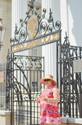 Woman in a pink floral sundress and wide-brim hat standing by an ornate black wrought-iron gate with a gold harp emblem and white-columned historic building in the background.