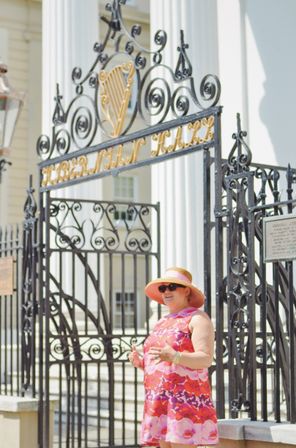 Woman in a pink floral sundress and wide-brim hat standing by an ornate black wrought-iron gate with a gold harp emblem and white-columned historic building in the background.