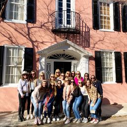 Group of friends posing in front of a pastel pink historic townhouse with black shutters, white pedimented doorway and small iron balcony on a sunny street.