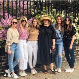 Six smiling women in casual spring outfits (denim, white jeans, straw hat, sunglasses) posing on a city sidewalk in front of a black iron fence covered with pink and white azalea blooms.