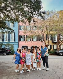Eight friends in bright summer dresses and sneakers smiling on a tree-lined street in front of pastel historic row houses (blue, pink, yellow) in Charleston, South Carolina.