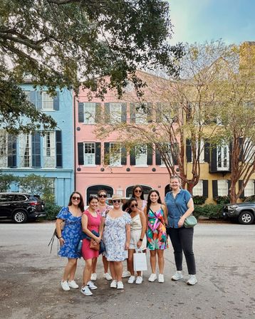 Eight friends in bright summer dresses and sneakers smiling on a tree-lined street in front of pastel historic row houses (blue, pink, yellow) in Charleston, South Carolina.