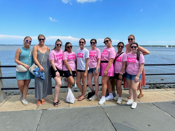 Group of friends in casual summer outfits, many wearing matching pink 'Groovy & Boozy' shirts, posing on a waterfront promenade with a calm bay, sailboats, and blue sky in the background.