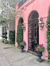 Charming pink stucco façade with arched wrought-iron gate, wall lanterns, potted flower urns and a blooming street tree lining a city sidewalk.
