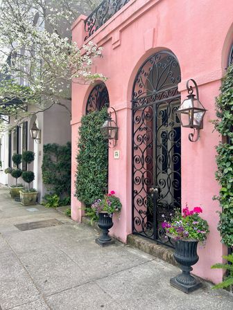 Charming pink stucco façade with arched wrought-iron gate, wall lanterns, potted flower urns and a blooming street tree lining a city sidewalk.
