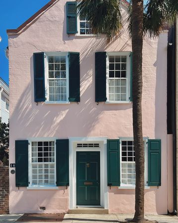 Charming pastel pink two-story house with dark green shutters and door, a tall palm tree casting sunlit shadows against a clear blue sky